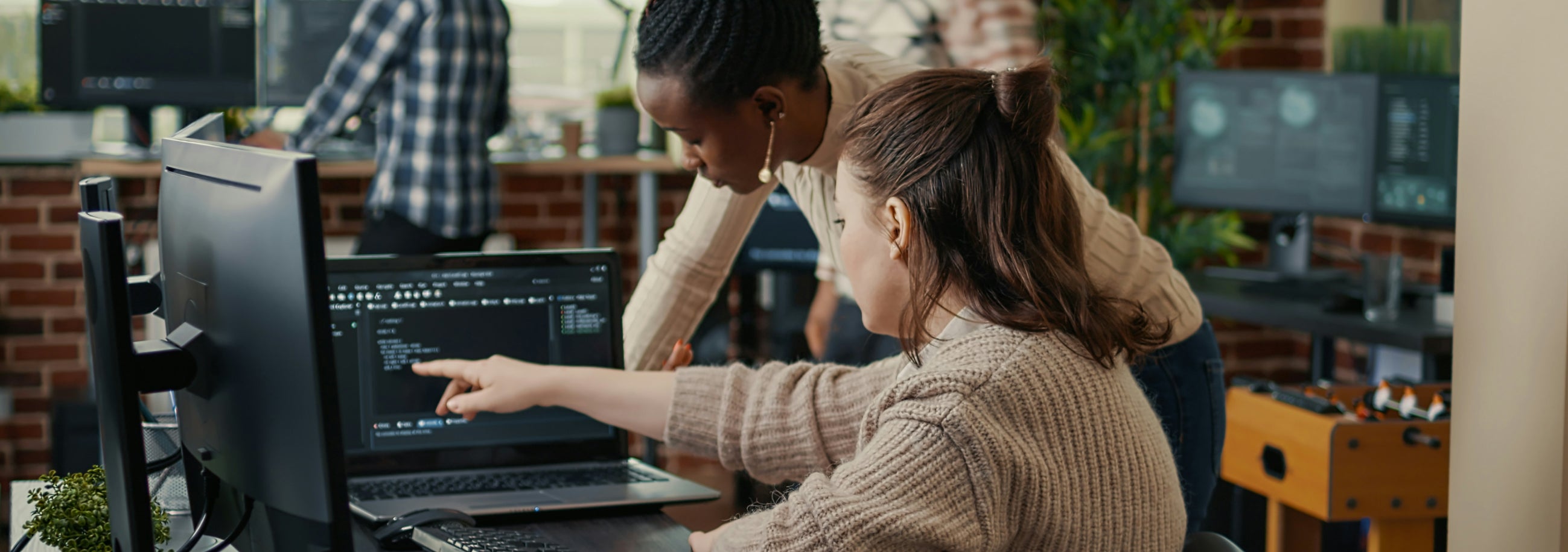 Two people at a desk with a laptop displaying code; one is pointing at the screen.