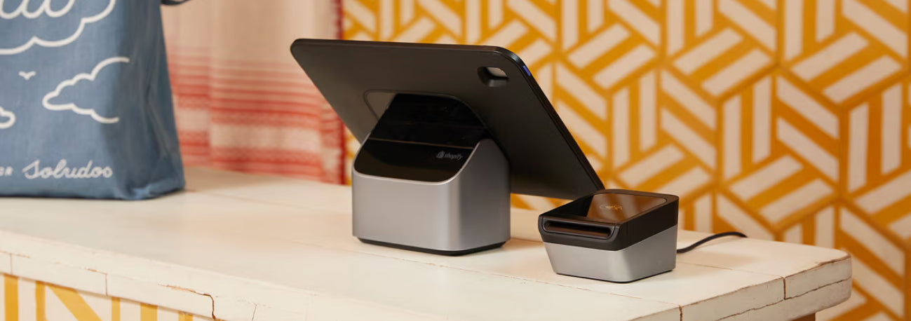 Shopify POS hardware set on a white rustic counter against a vibrant geometric-patterned backdrop, featuring a tablet display and card reader.